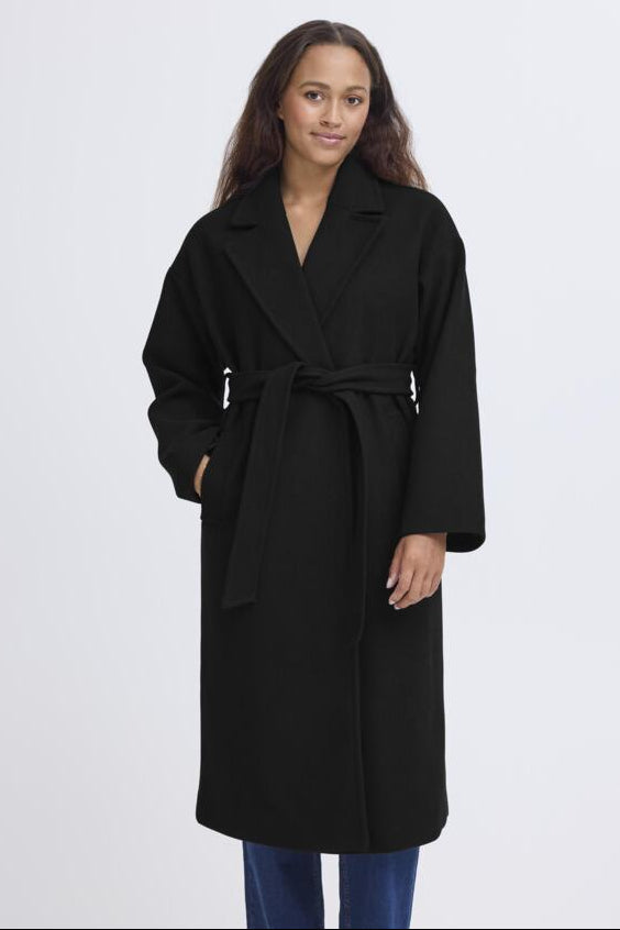 A woman modeling a long black belted coat against a plain light gray studio background.