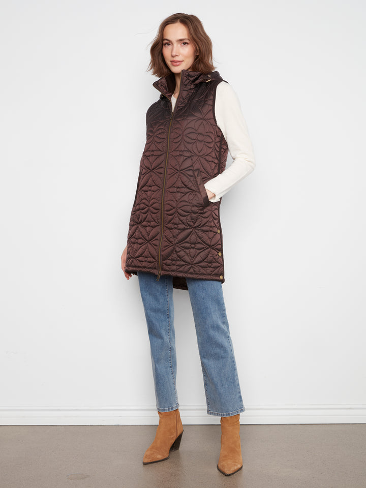 A woman modeling a dark brown quilted sleeveless puffer vest over a white long-sleeve top, blue jeans, and tan ankle boots, standing against a plain white wall.