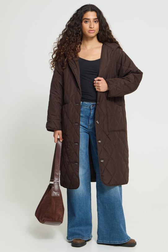 A woman stands against a light gray studio background wearing a long dark brown quilted coat over a black top and blue wide-leg jeans, while holding a brown shoulder bag.