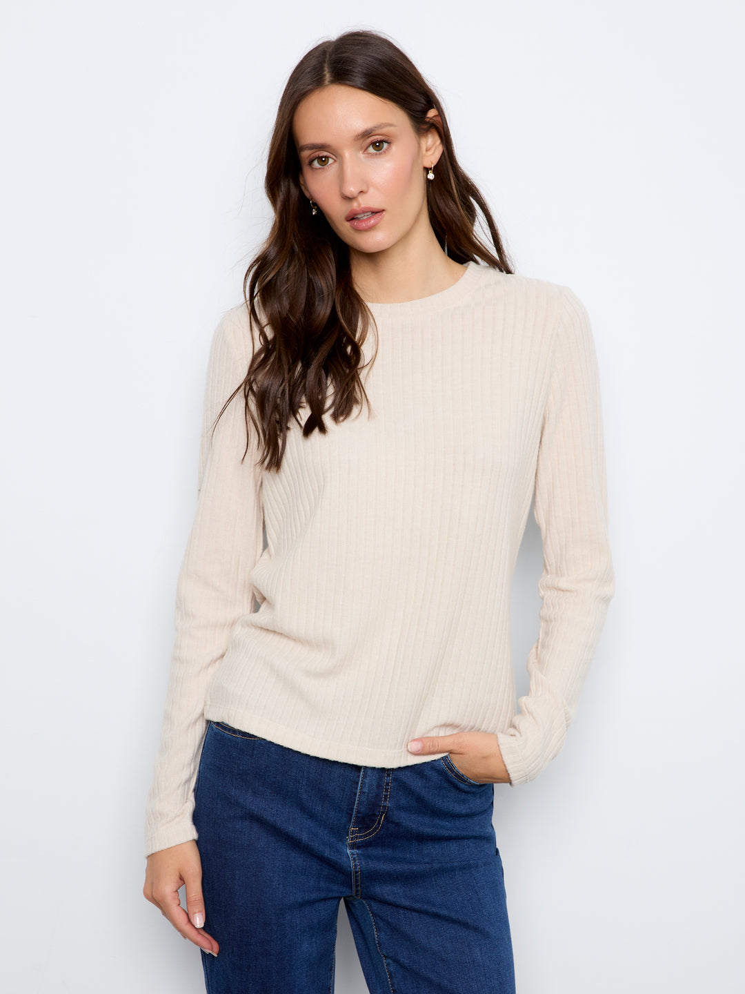A woman wearing a cream ribbed long-sleeve knit sweater and dark blue jeans, standing against a plain light gray studio background.