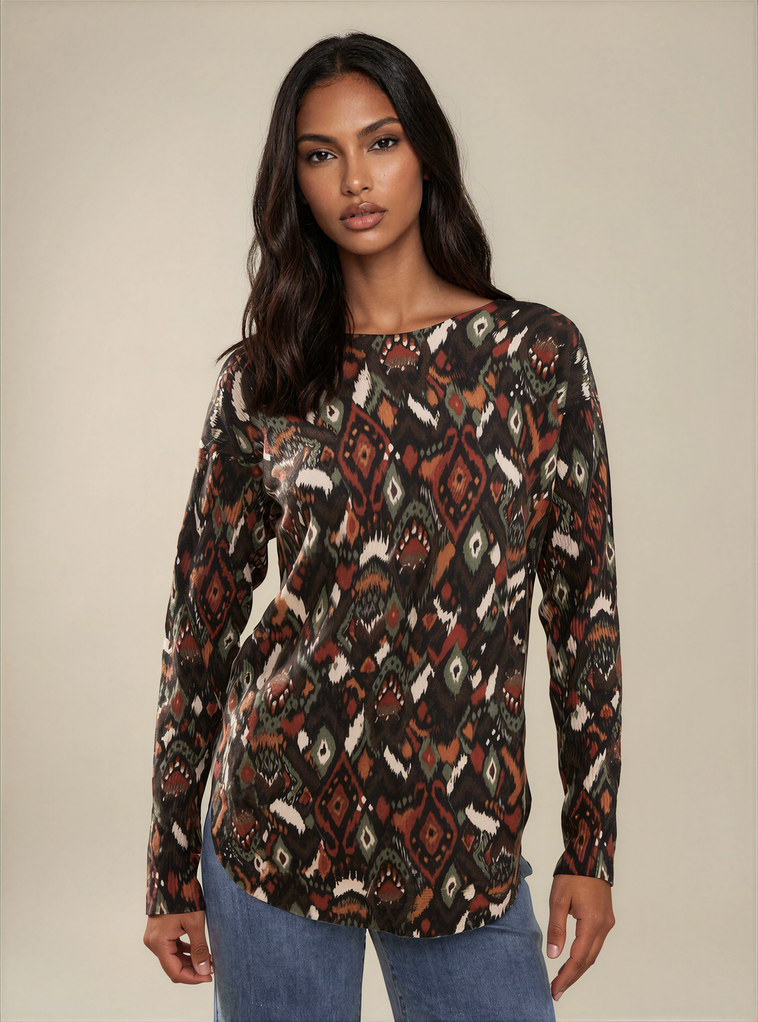 A woman modeling a long-sleeve bohemian-style patterned top with geometric tribal motifs in brown, rust, beige, and black, paired with blue jeans against a solid beige studio background.