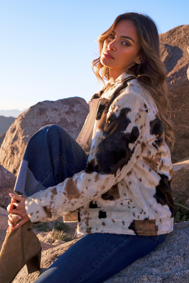 A woman sitting outdoors on a rock in a desert landscape, wearing a fuzzy cropped jacket with a black, brown, and beige cow-print pattern, blue jeans, and brown ankle boots.