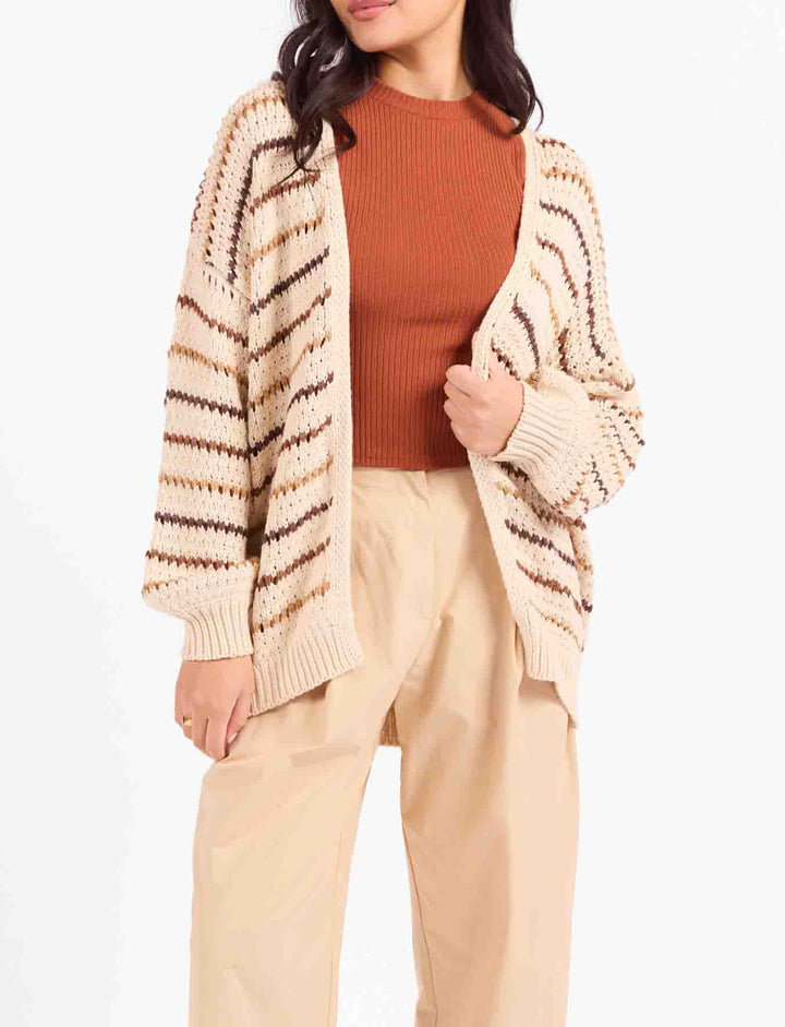 A woman modeling a beige and brown striped knit cardigan over a rust-orange ribbed top, paired with beige cropped pants and tan loafers, against a plain white studio background.
