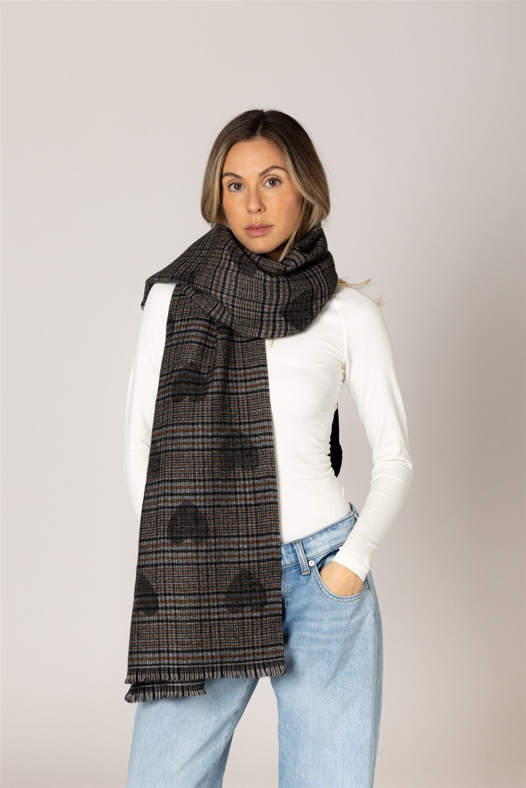 A woman modeling a dark gray and black plaid scarf with a subtle cloud pattern, styled over a white long-sleeve top and light blue jeans against a solid light gray background.