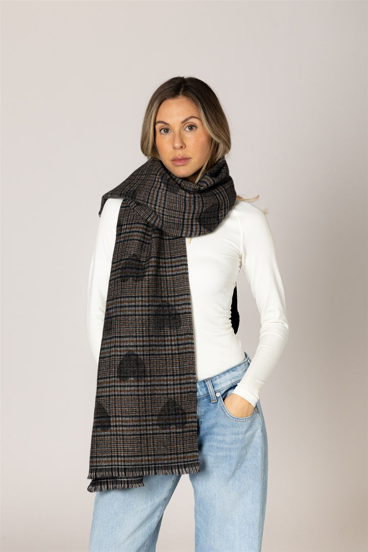 A woman modeling a dark gray and black plaid scarf with a subtle cloud pattern, styled over a white long-sleeve top and light blue jeans against a solid light gray background.