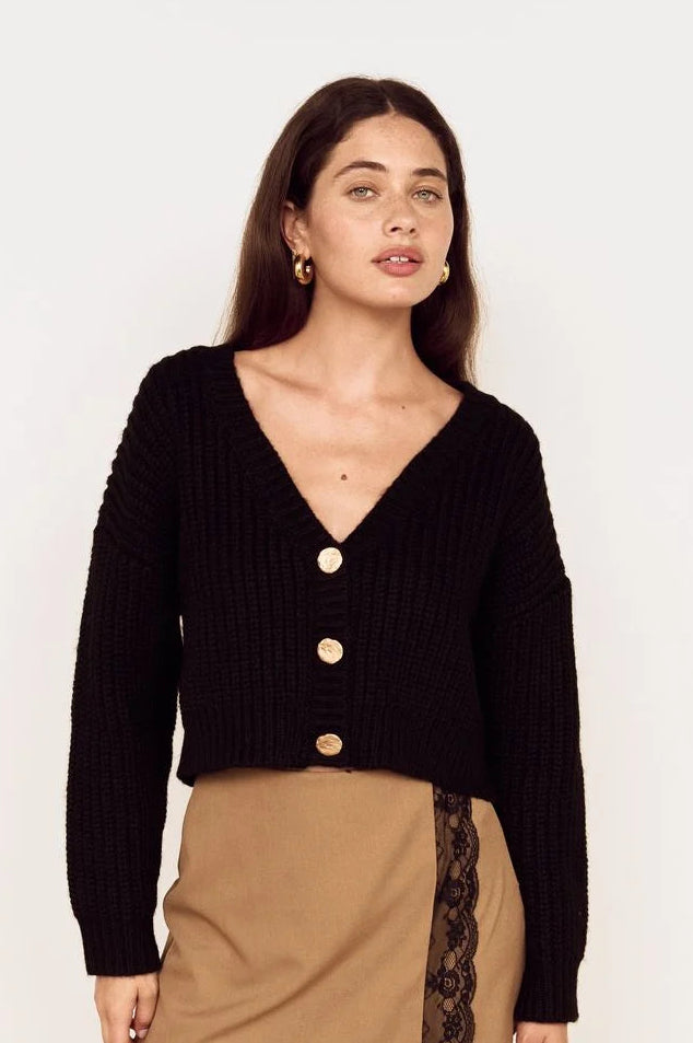 A woman modeling a black ribbed knit cardigan with three gold buttons, paired with a tan wrap skirt featuring black lace trim, black tights, and gold hoop earrings against a plain white background.
