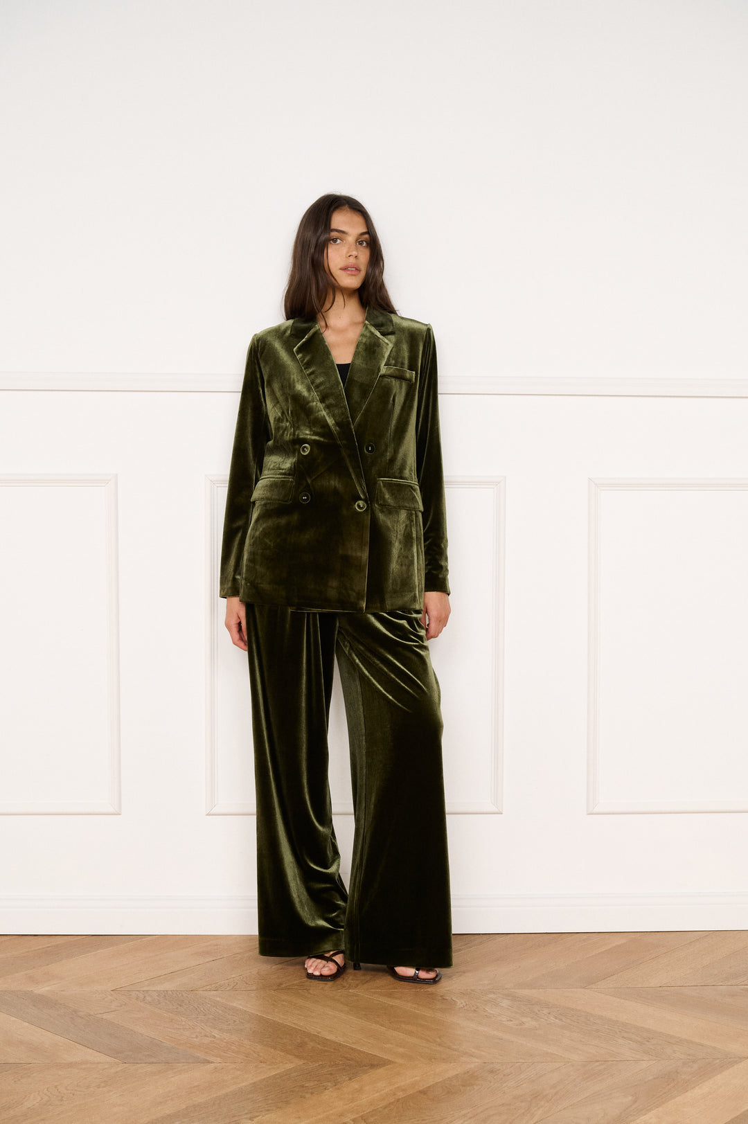 Woman wearing a green velvet suit standing in a room with wooden flooring and white walls.