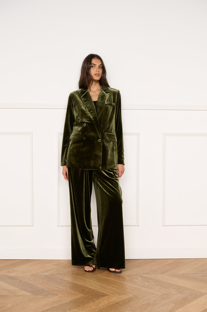 Woman wearing a green velvet suit standing in a room with wooden flooring and white walls.