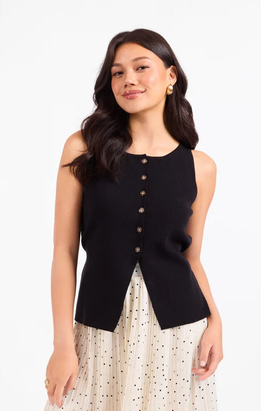 A woman modeling a sleeveless black knit vest with a button-down front, paired with a cream polka-dot skirt, against a plain white studio background.