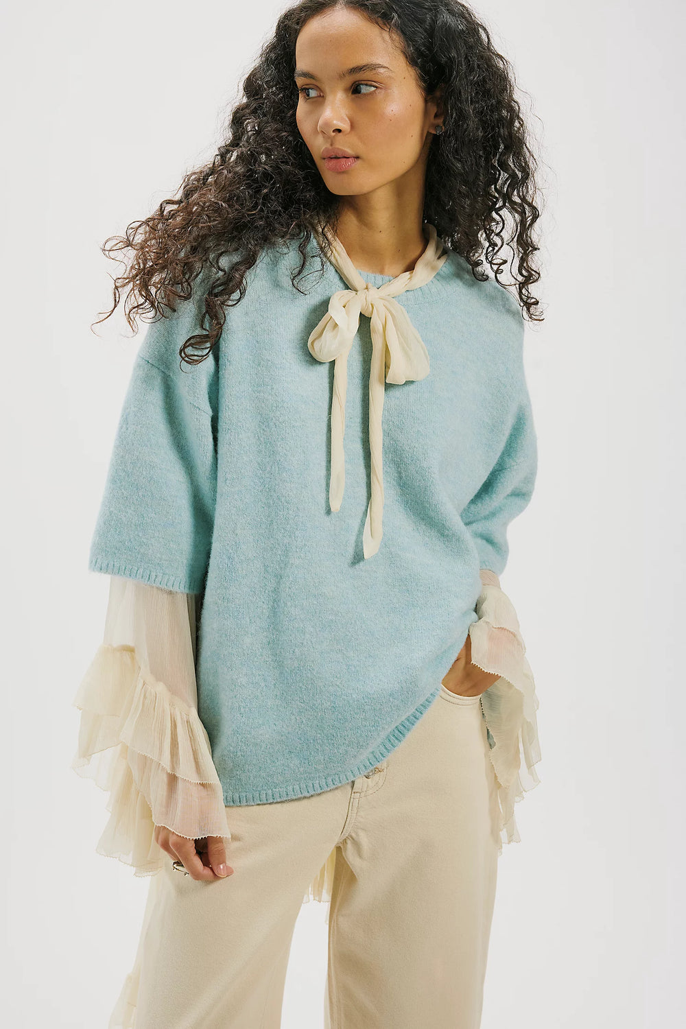 Woman wearing a light blue sweater with a beige bow tie and beige pants on a white background