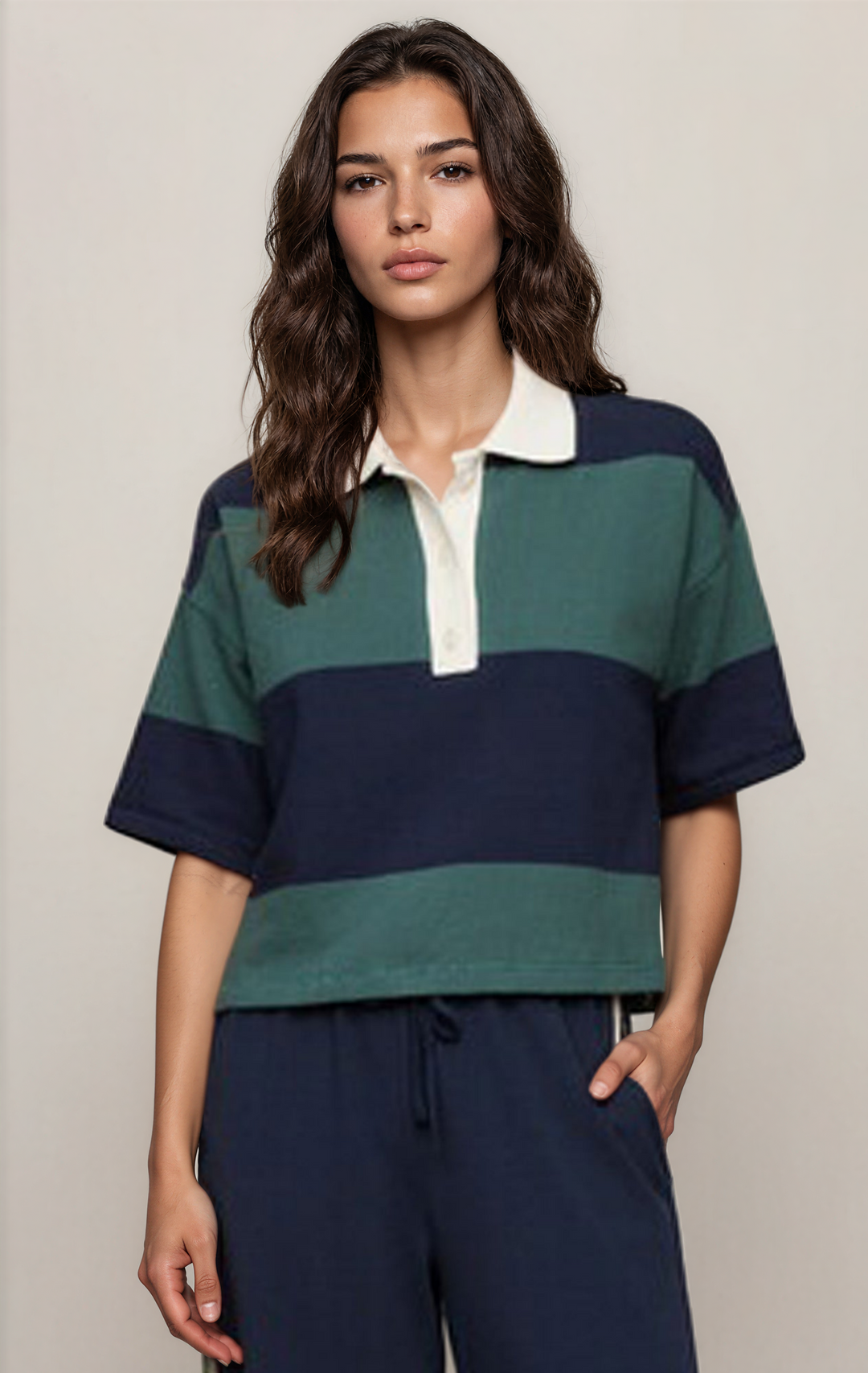 A woman modeling a short-sleeve rugby-style polo shirt with horizontal stripes in dark navy, forest green, and cream, paired with navy drawstring pants against a plain light beige studio background.