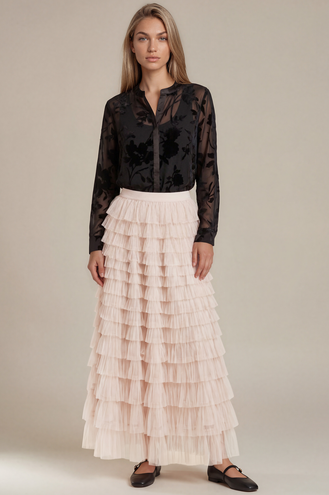 A model stands against a plain beige studio background wearing a sheer black long-sleeve blouse and a high-waisted, multi-tiered light pink pleated maxi skirt.