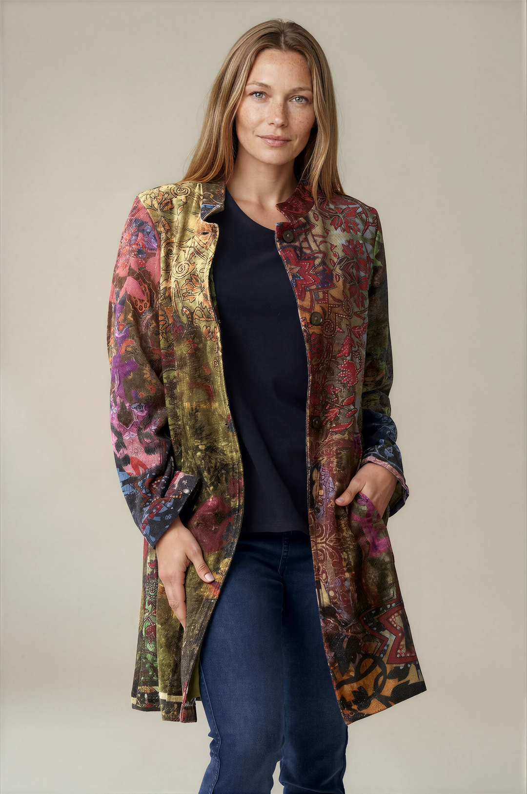 A woman modeling a long, open-front coat with a richly patterned multicolor exterior featuring gold, green, red, and purple tones, worn over a black top and blue jeans against a plain beige studio background.