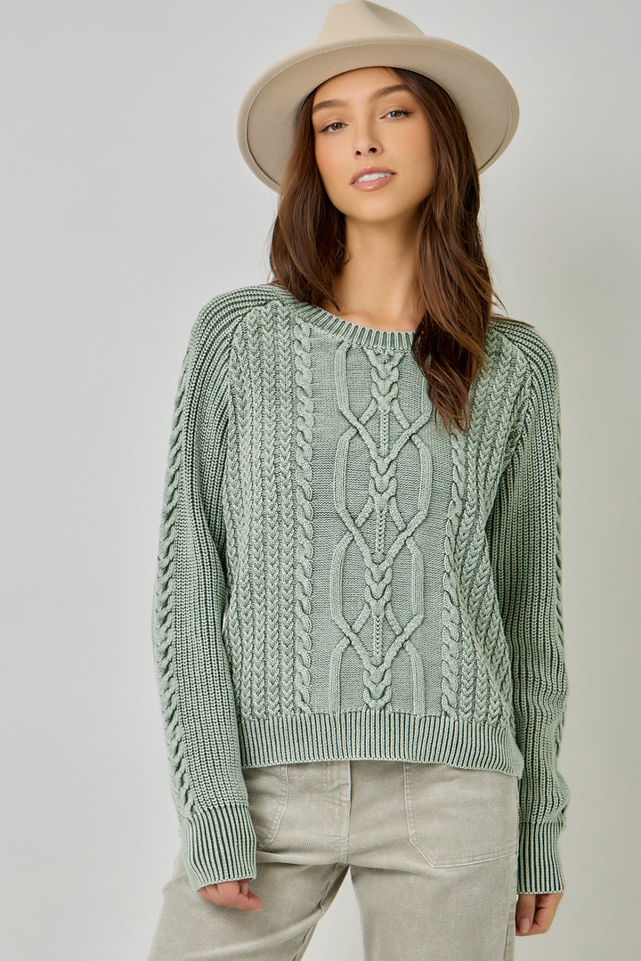 A woman wearing a sage green cable-knit sweater, light beige pants, and a beige wide-brim hat, photographed against a neutral light gray studio background.