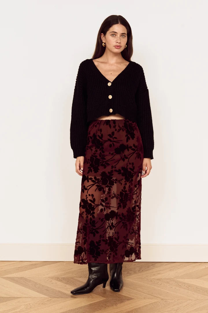 A woman stands in a studio wearing a black cropped cardigan with gold buttons and a dark burgundy floral midi skirt with black boots.
