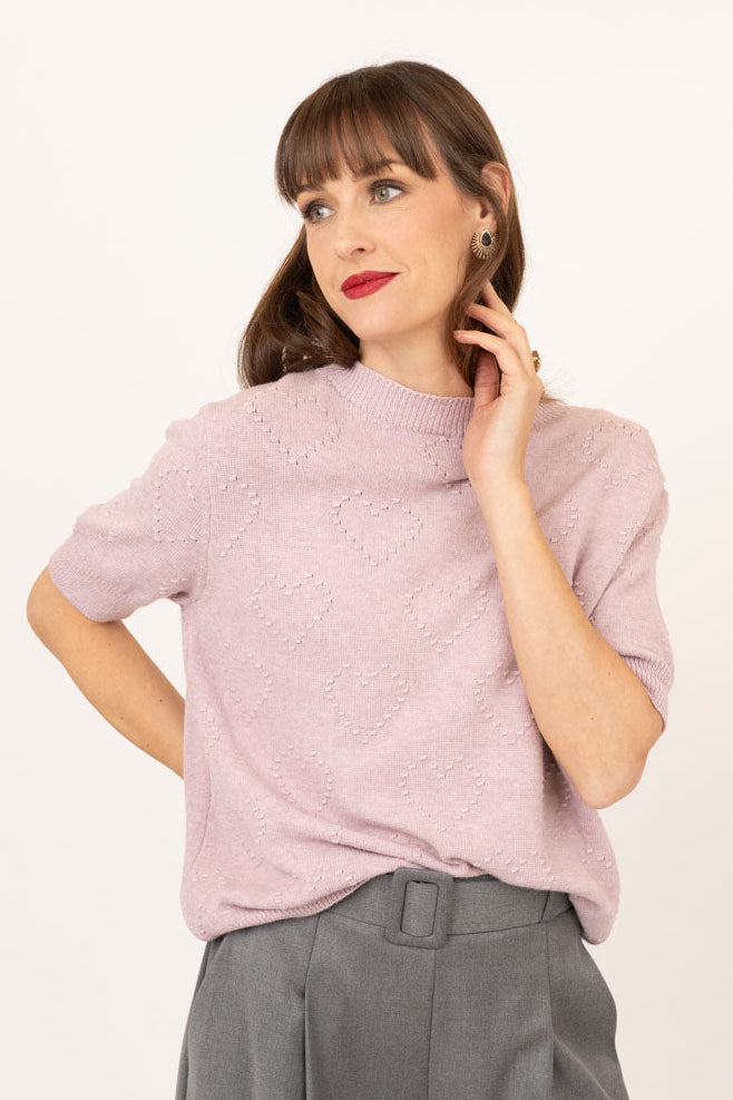 A woman modeling a short-sleeve knit top in a dusty pink color, paired with high-waisted gray wide-leg trousers, against a plain off-white studio background.
