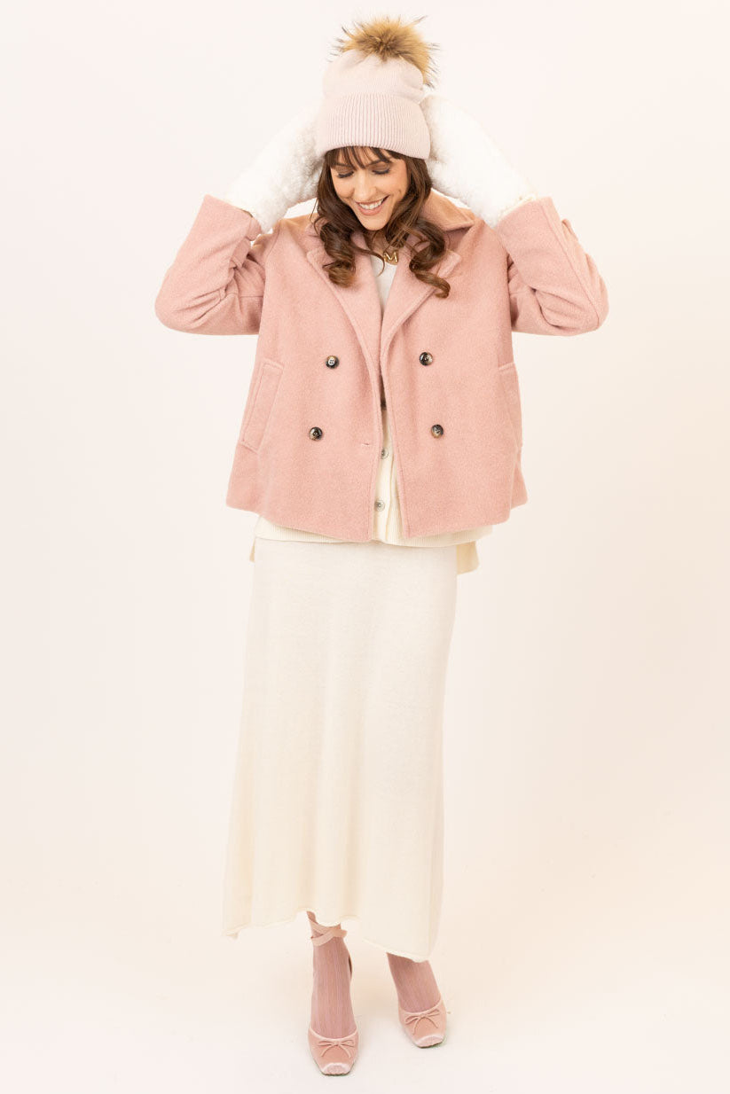 A woman modeling a light pink double-breasted jacket, cream maxi skirt, matching pink knit beanie with a brown pom-pom, and pink pointed-toe heels against a plain off-white studio background.