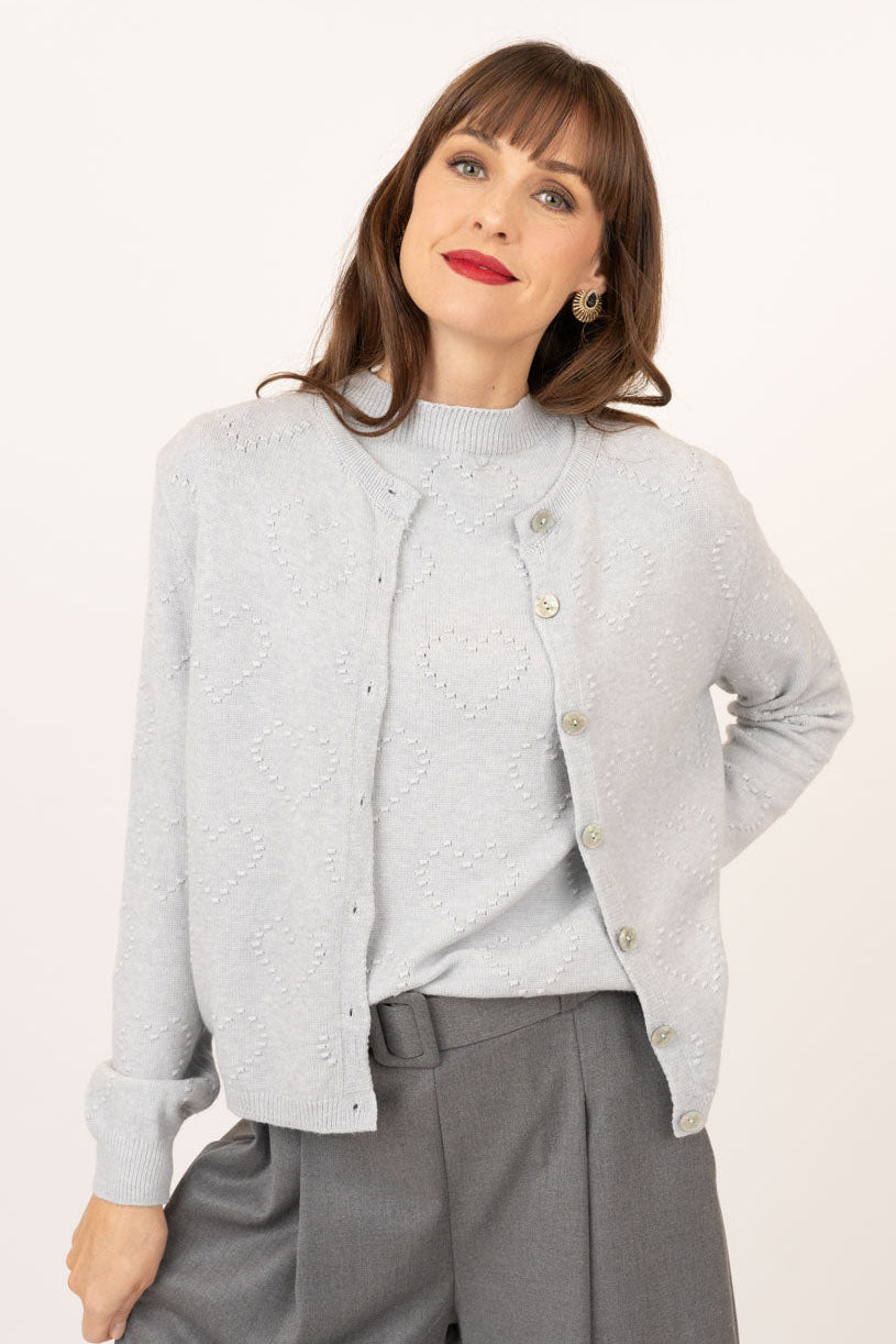 A woman modeling a light gray textured knit cardigan with gold buttons over a matching gray top and gray trousers, set against a plain off-white studio background.