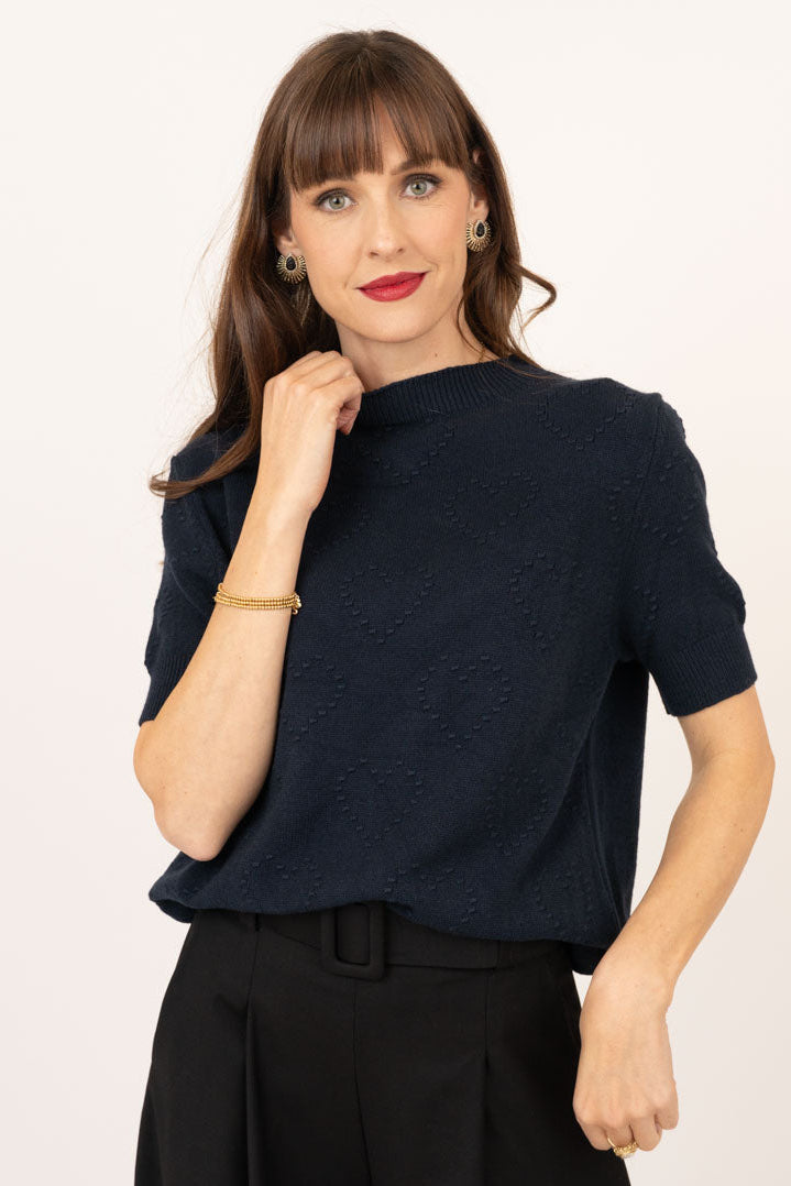 A woman modeling a short-sleeve textured knit top in dark navy blue, paired with black wide-leg trousers, against a plain off-white studio background.