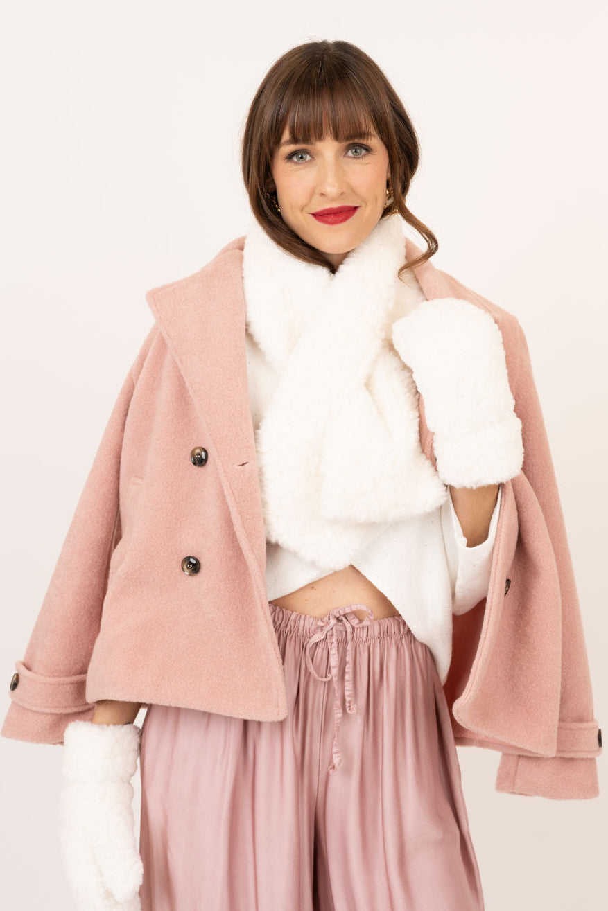 A woman modeling a light pink double-breasted coat with white faux-fur trim, a white cropped top, white mittens, and dusty pink wide-leg trousers against a plain off-white background.