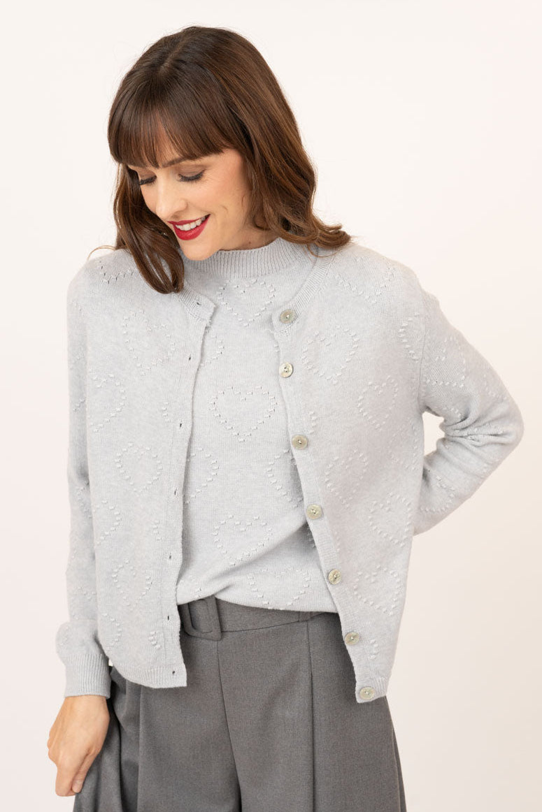 A woman wearing a light gray textured knit cardigan with gold-tone buttons over a matching gray top and gray wide-leg trousers, photographed against a plain off-white studio background.
