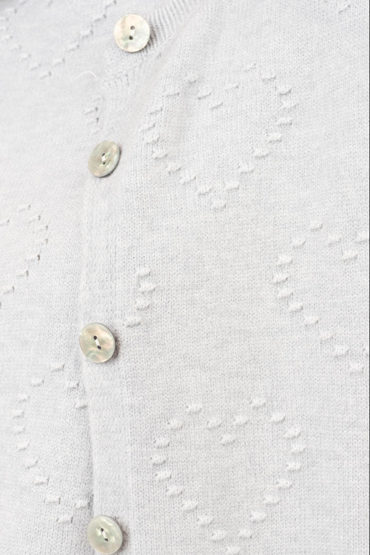 Close-up of a light gray knit cardigan featuring a textured heart pattern and a vertical row of cream-colored buttons.
