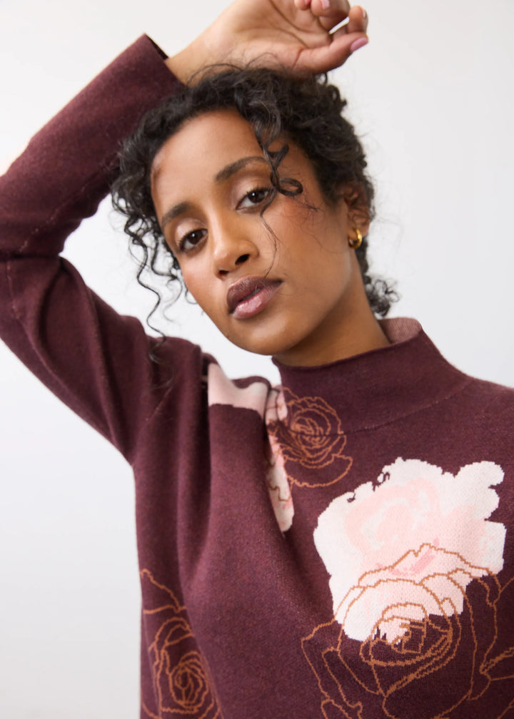 A woman wearing a burgundy mock-neck knit sweater with a large light pink rose applique and embroidered rose details, posing against a plain light gray background.