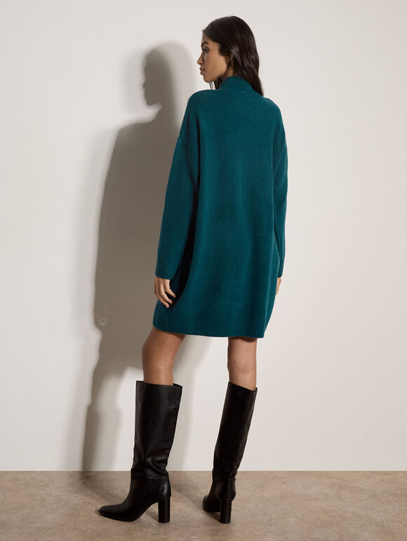 A woman stands with her back to the camera wearing a teal green knit sweater dress and black knee-high leather boots against a plain off-white wall.