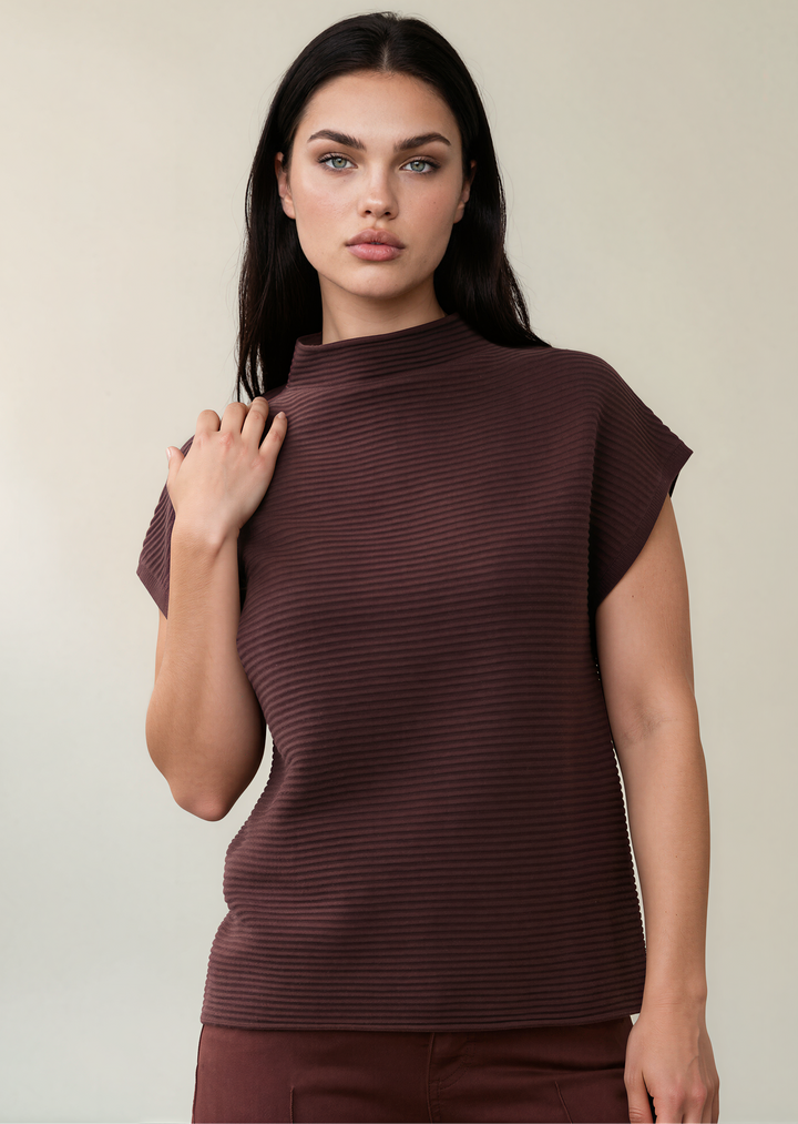 A woman modeling a ribbed, short-sleeve mock neck top in a deep plum-brown color, paired with matching brown pants, against a solid light beige background.