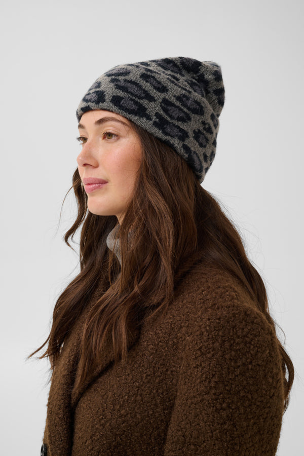 A woman in profile wearing a gray and black patterned knit beanie and a brown textured coat, photographed against a plain light gray studio background.