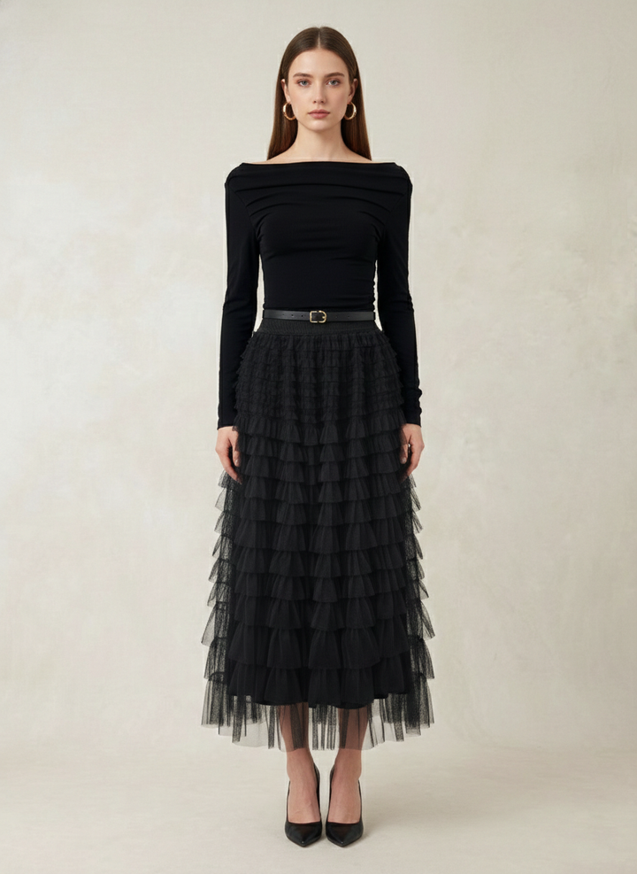 A model stands against a textured beige studio backdrop wearing a black off-the-shoulder long-sleeve top paired with a black tiered tulle midi skirt and a thin black belt with a gold buckle.