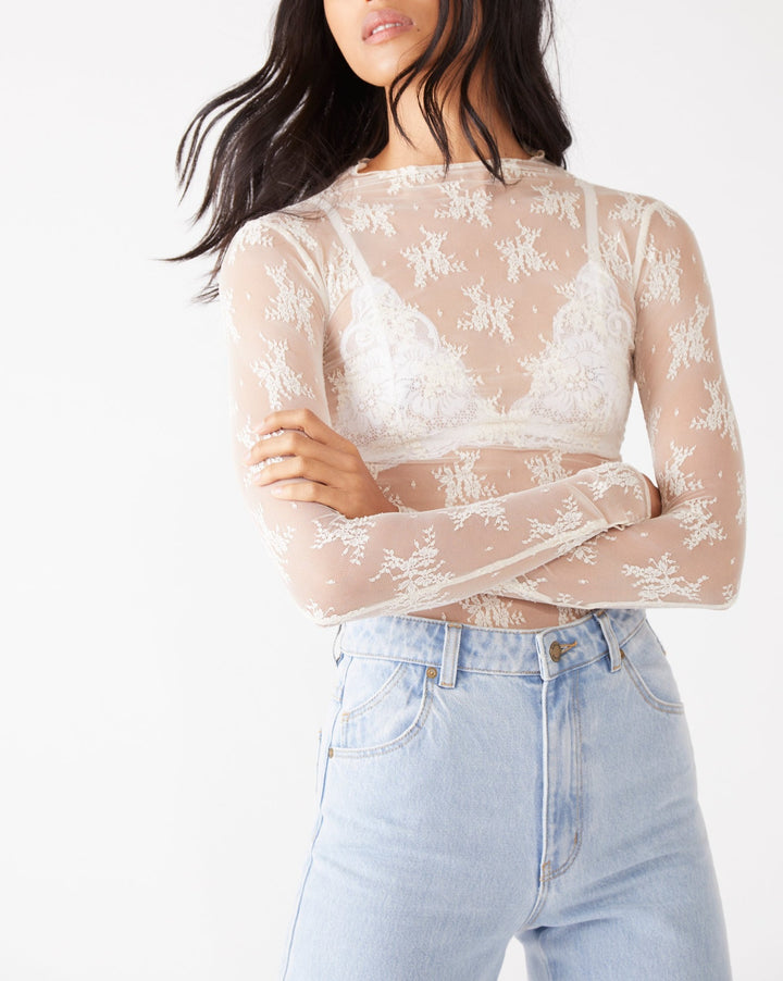 A woman wearing a sheer beige long-sleeve crop top with white floral lace embroidery over a white underwire bra, paired with light blue jeans, against a plain white background.