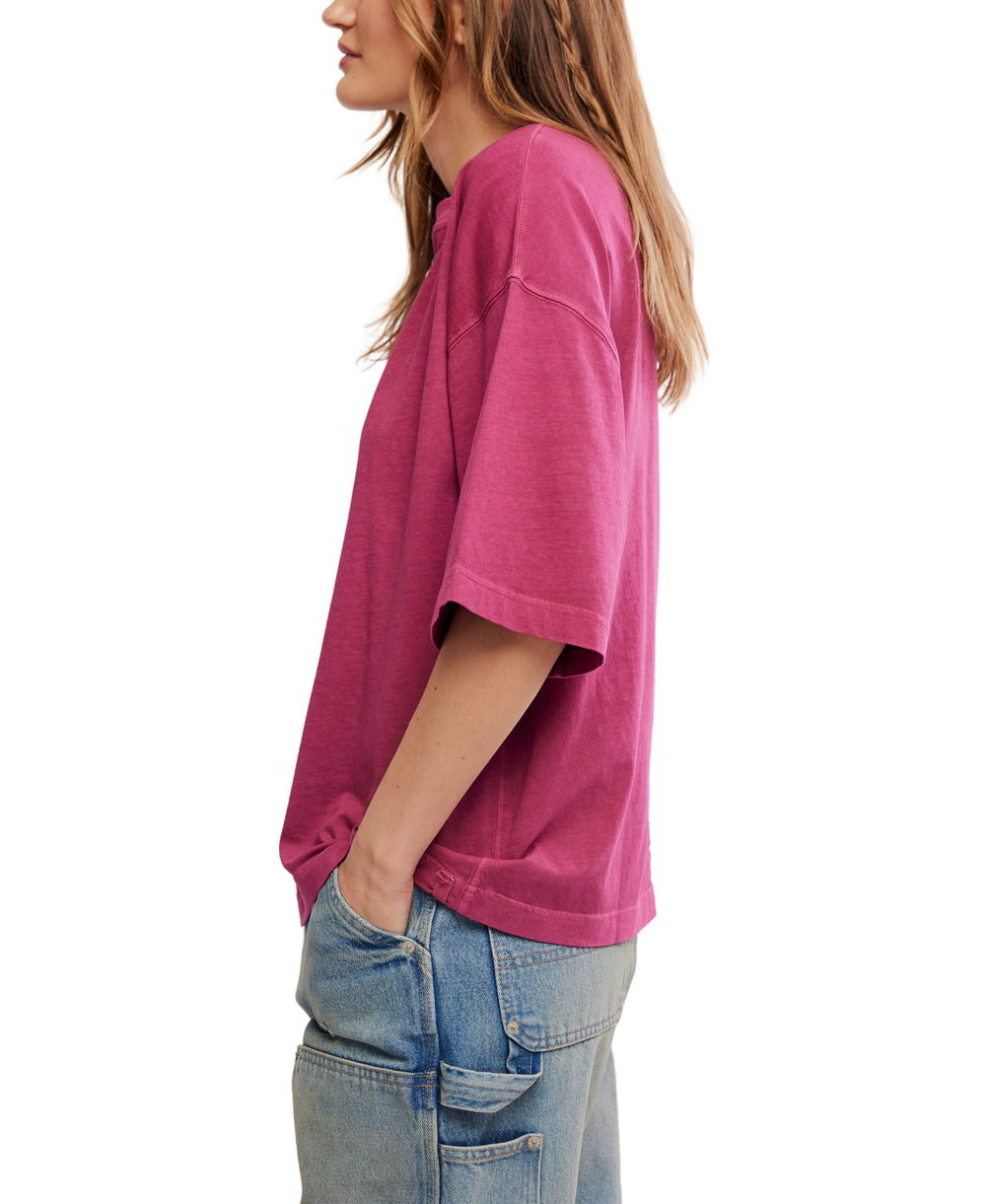 A woman shown in side profile wearing an oversized short-sleeve t-shirt in a washed magenta-pink color, paired with light-wash blue jeans, against a plain white background.