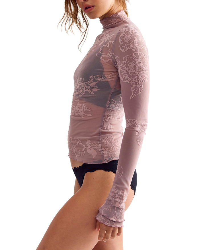 A woman wearing a sheer, long-sleeve turtleneck top in dusty mauve pink with white floral lace embroidery, paired with black lace-trimmed underwear, shown against a plain white background.