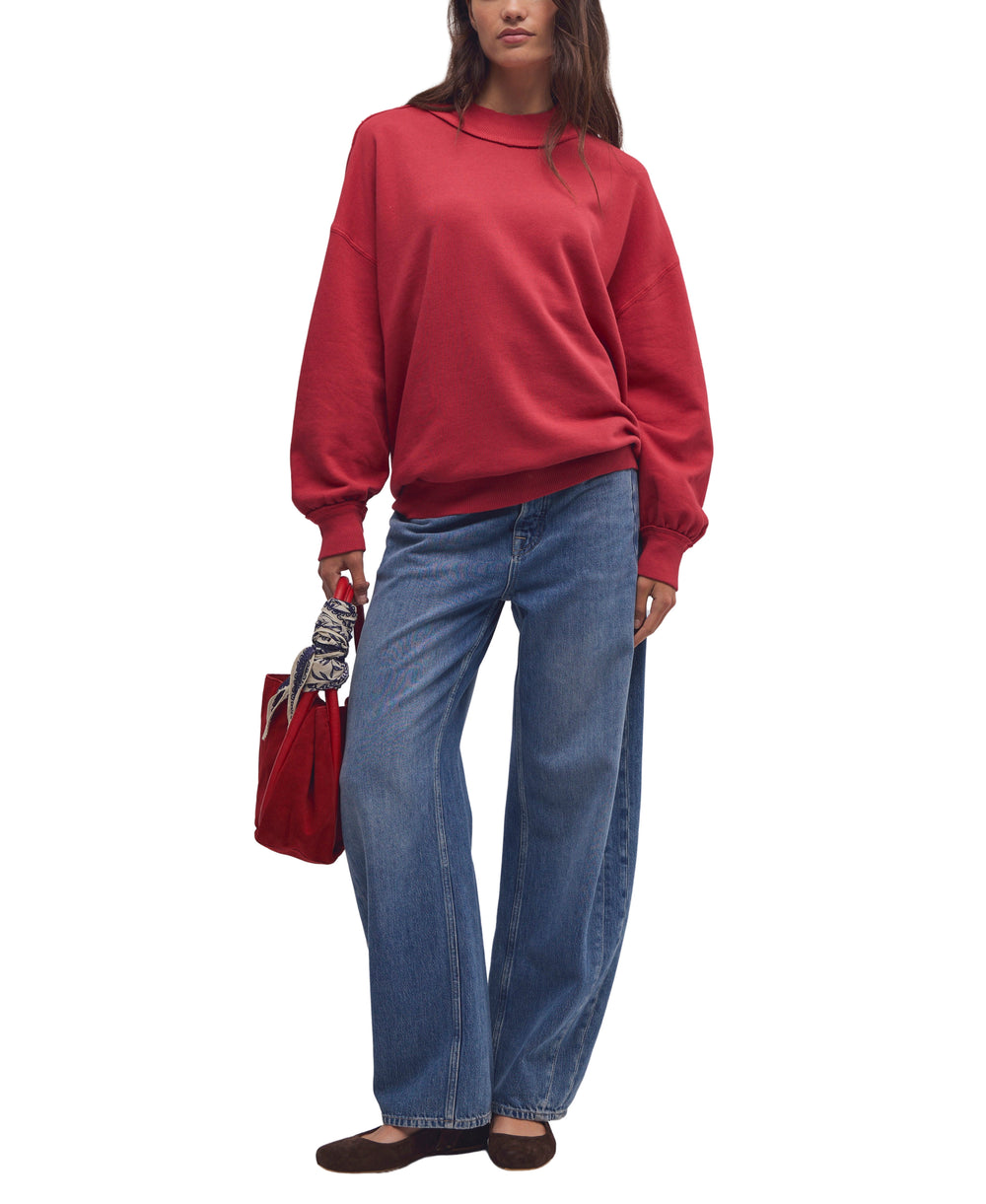 A model wearing a red crewneck sweatshirt and wide-leg blue jeans, holding a red handbag against a plain white background.