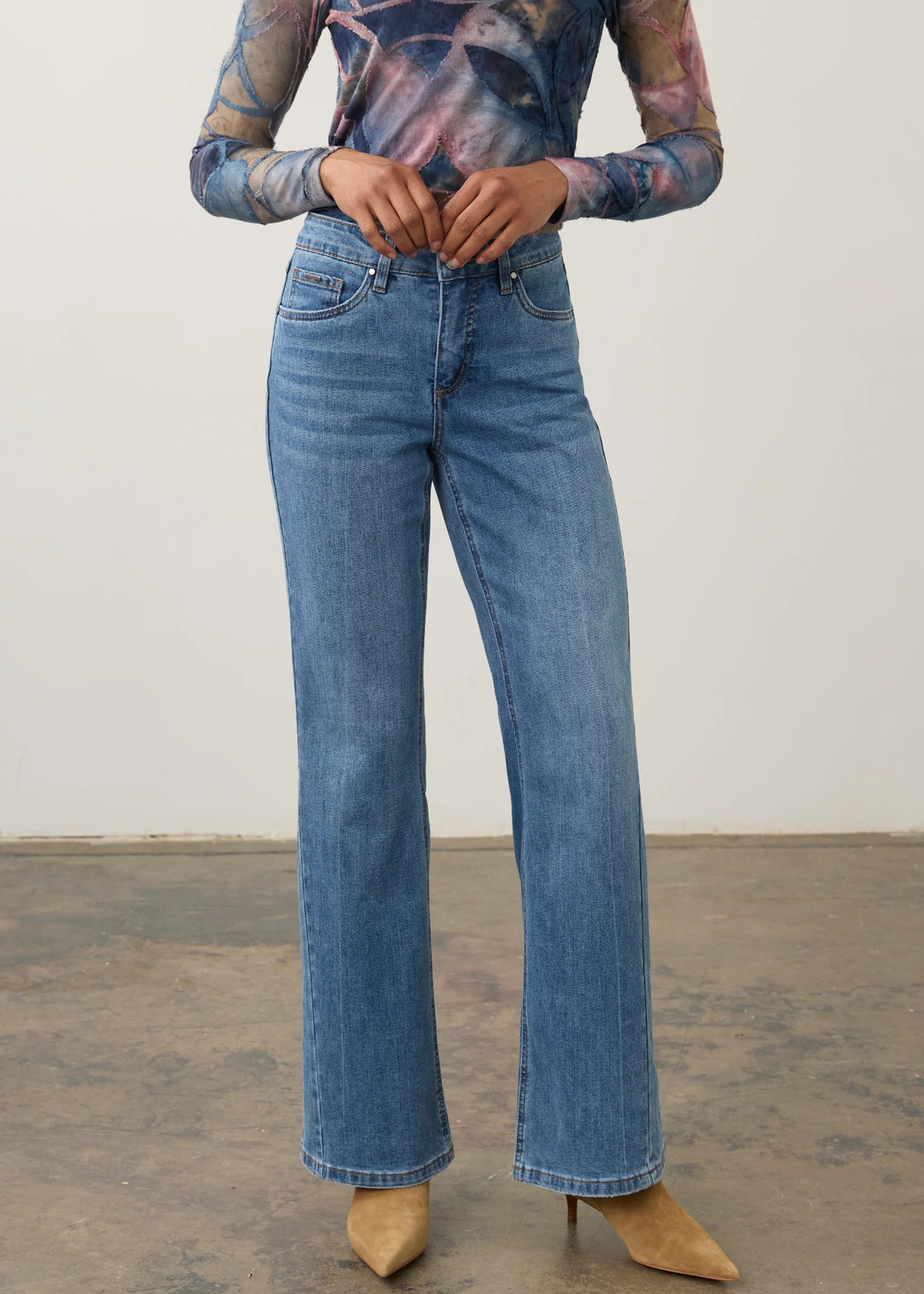 A model wearing high-waisted, wide-leg blue denim jeans with a floral long-sleeve top and tan pointed-toe heels, standing against a plain off-white wall and concrete floor.