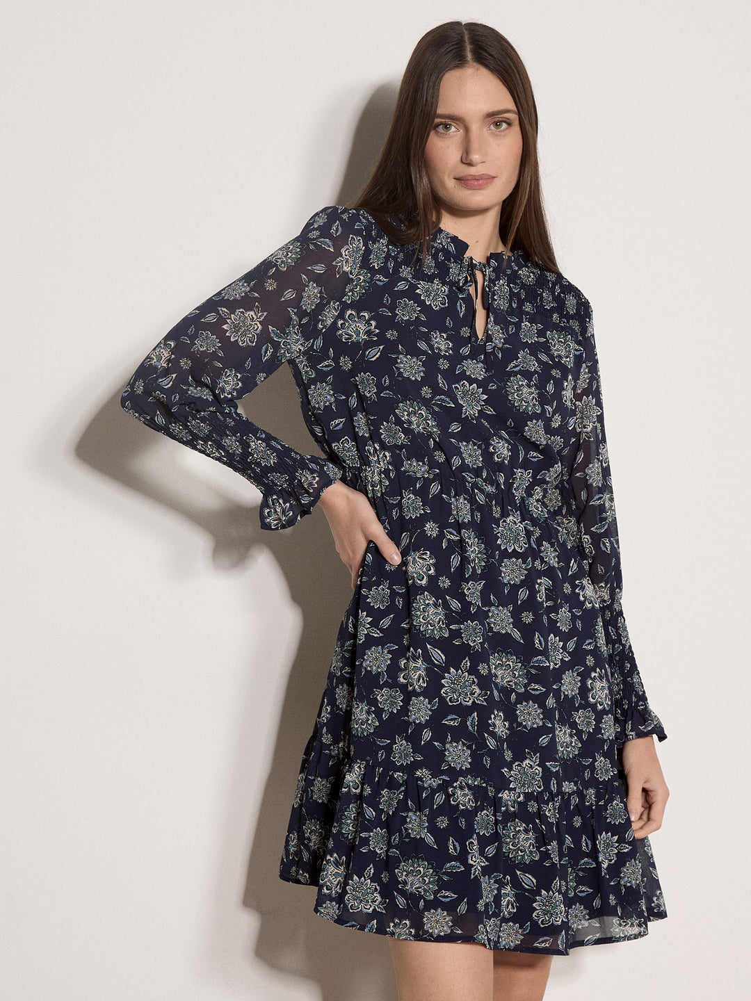 A woman modeling a navy blue long-sleeve dress with a white floral print, featuring a tie-neck detail and a flared skirt, set against a plain off-white background.