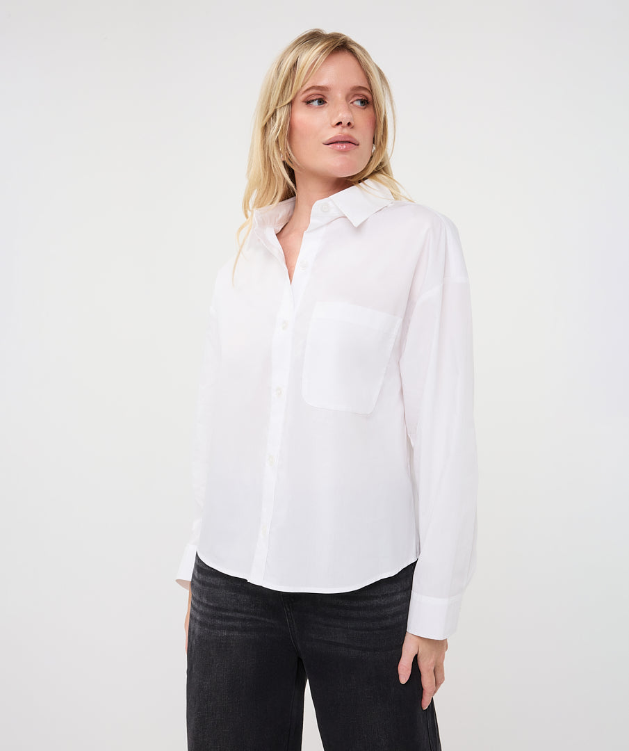 A blonde woman wearing a white long-sleeve button-up shirt with a chest pocket and black pants, standing against a plain white studio background.