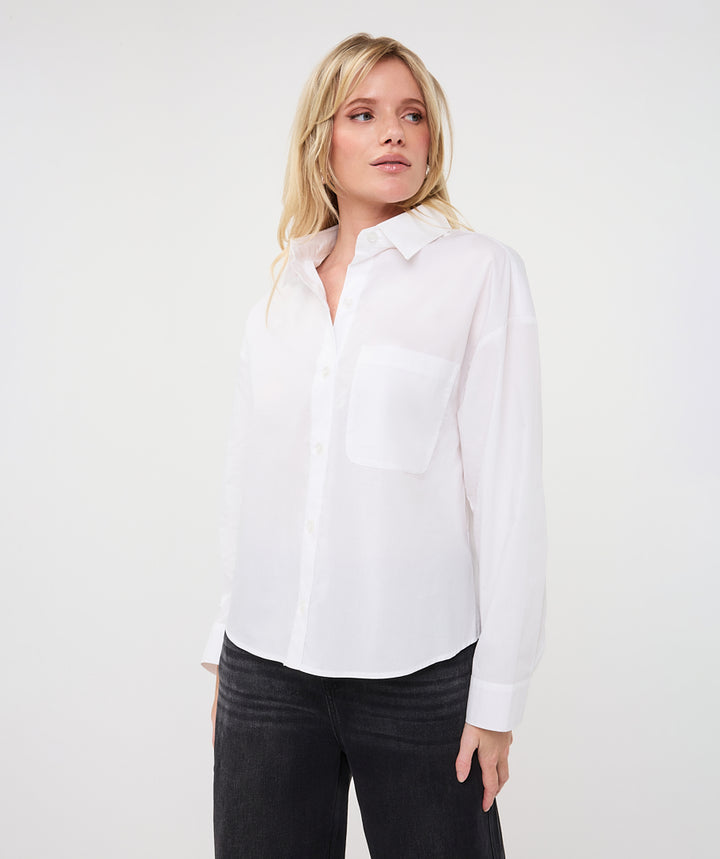 A blonde woman wearing a white long-sleeve button-up shirt with a chest pocket and black pants, standing against a plain white studio background.