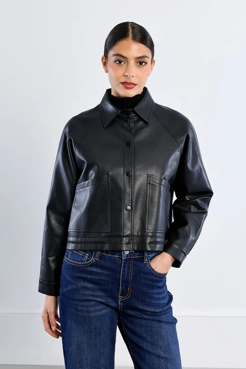 A woman modeling a black leather cropped jacket with a high collar and front button closure, paired with blue jeans, against a light gray studio background.