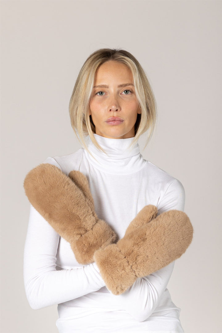 A woman with blonde hair wearing a white turtleneck sweater and fluffy tan faux-fur arm warmers, posing with her arms crossed against a solid light beige background.