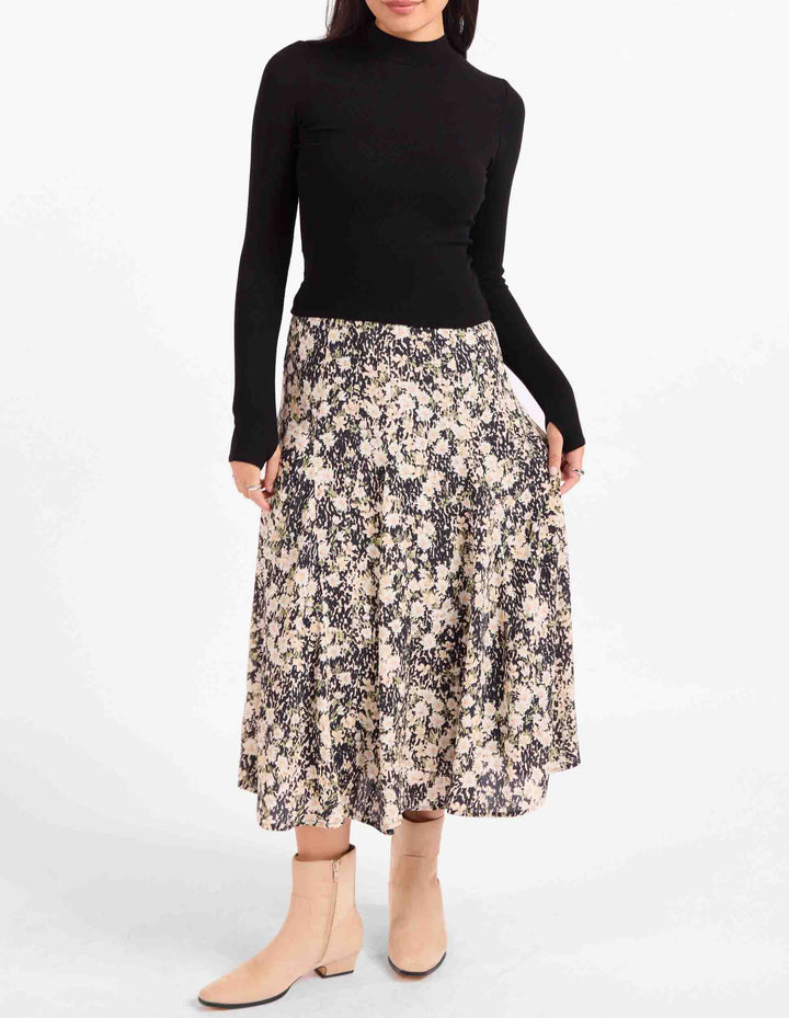 A model wearing a black long-sleeve mock neck top and a knee-length floral midi skirt with beige ankle boots, shot against a plain white studio background.