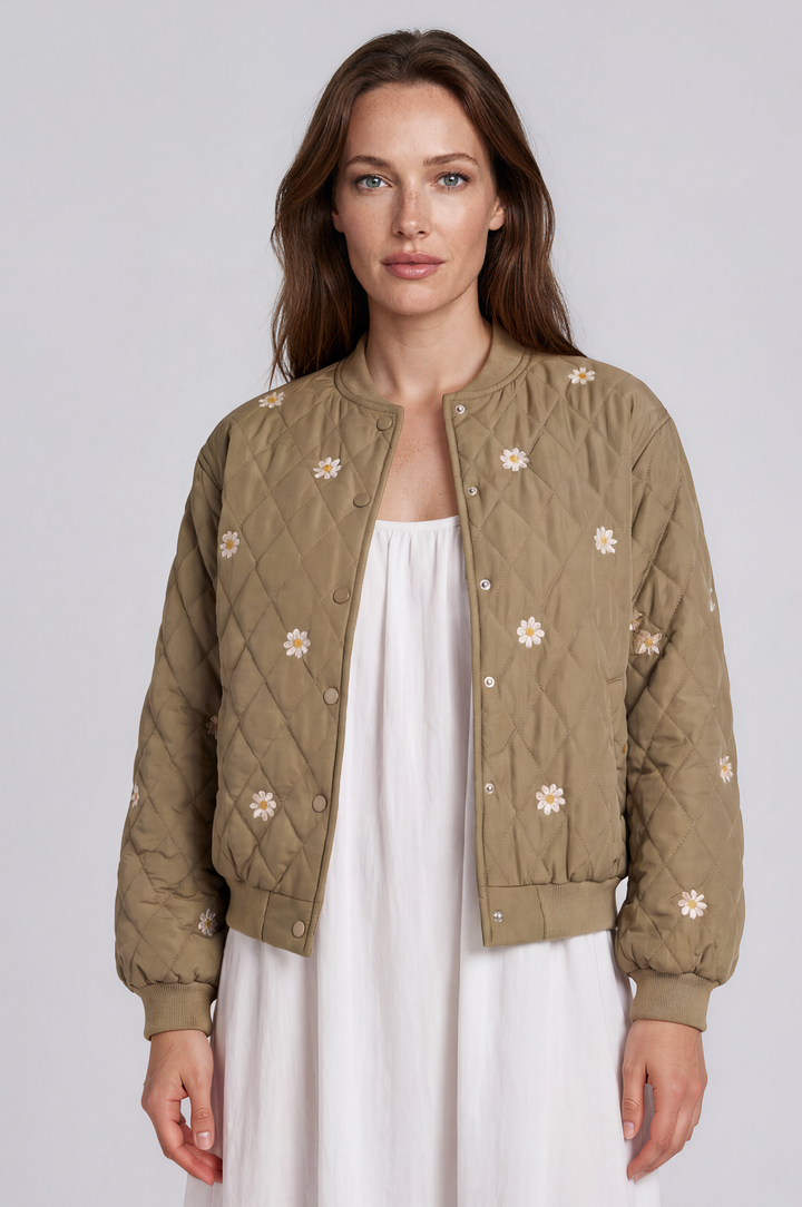 A woman wearing a quilted olive green bomber jacket with white floral embroidery over a white dress, photographed against a neutral light gray studio background.