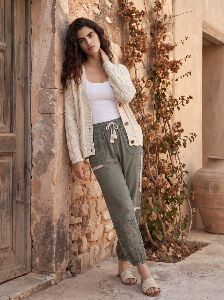Woman in a white cardigan and green pants standing against a stone wall with a plant.