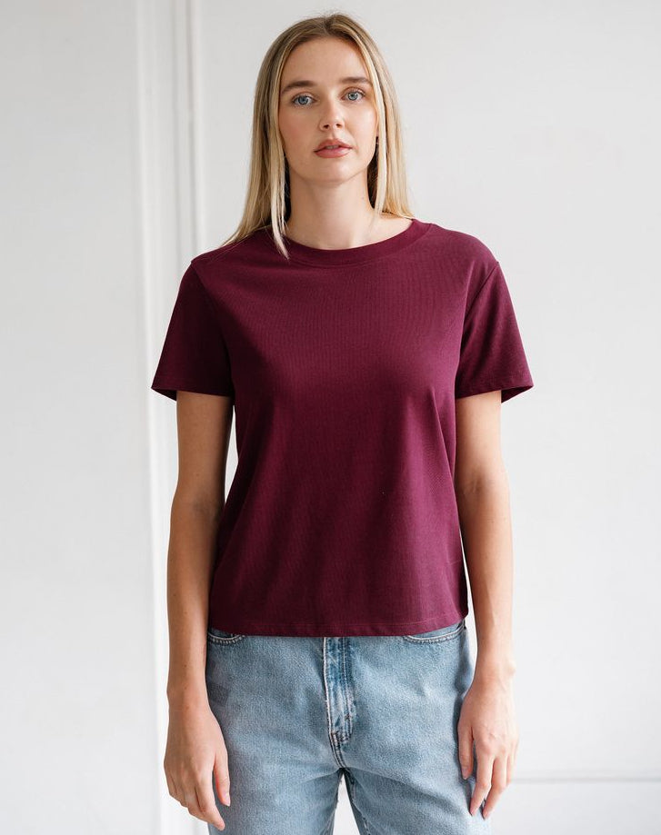A woman modeling a short-sleeve crewneck t-shirt in burgundy, paired with light-wash blue jeans, standing against a white wall.