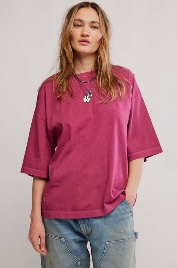 A woman wearing an oversized magenta pink t-shirt with three-quarter sleeves and light-wash blue jeans, standing against a neutral light gray background.