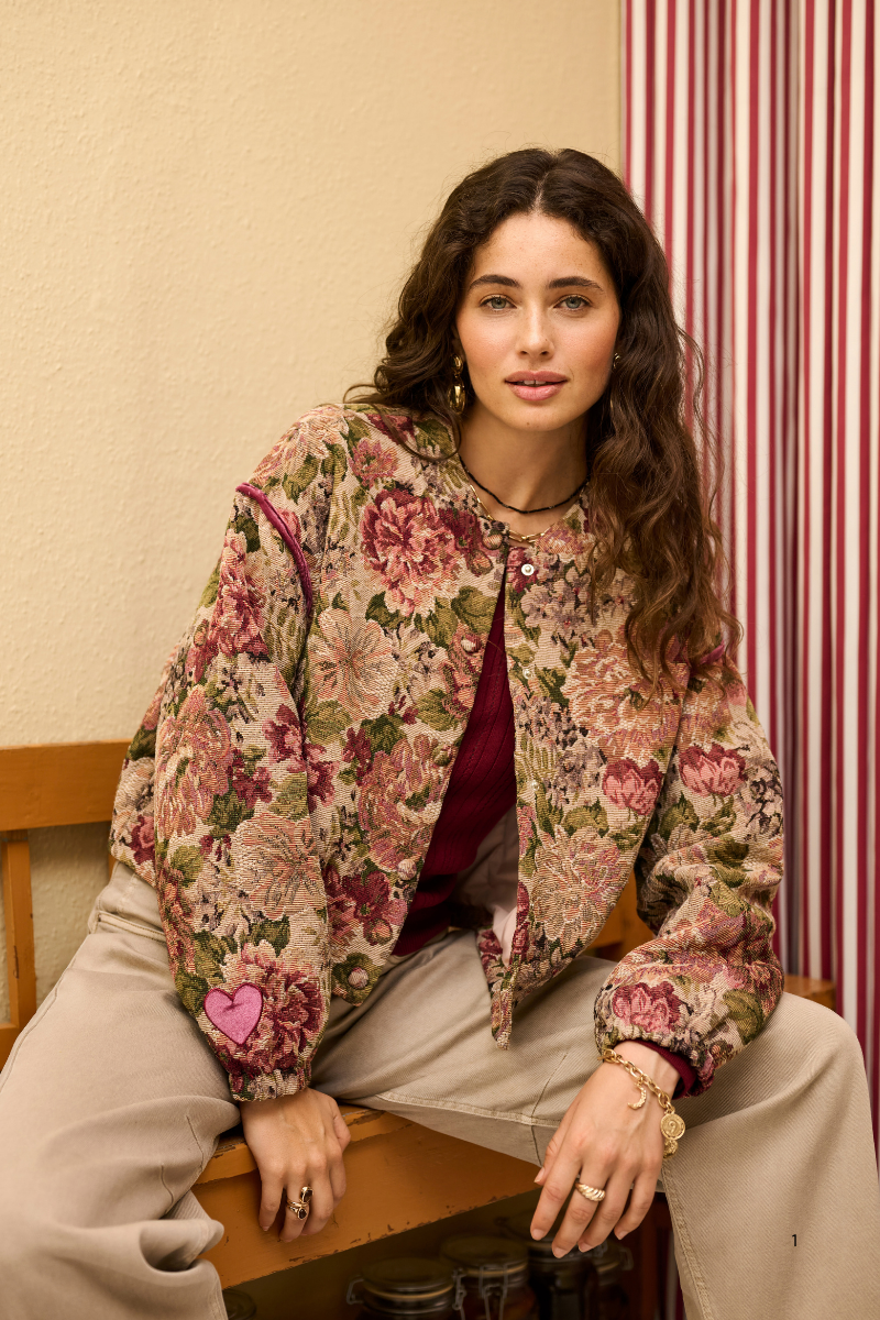 A woman sits on a wooden bench wearing a floral quilted jacket with pink roses and green leaves over a burgundy top and beige pants, set against a cream wall and red-and-white striped curtain.