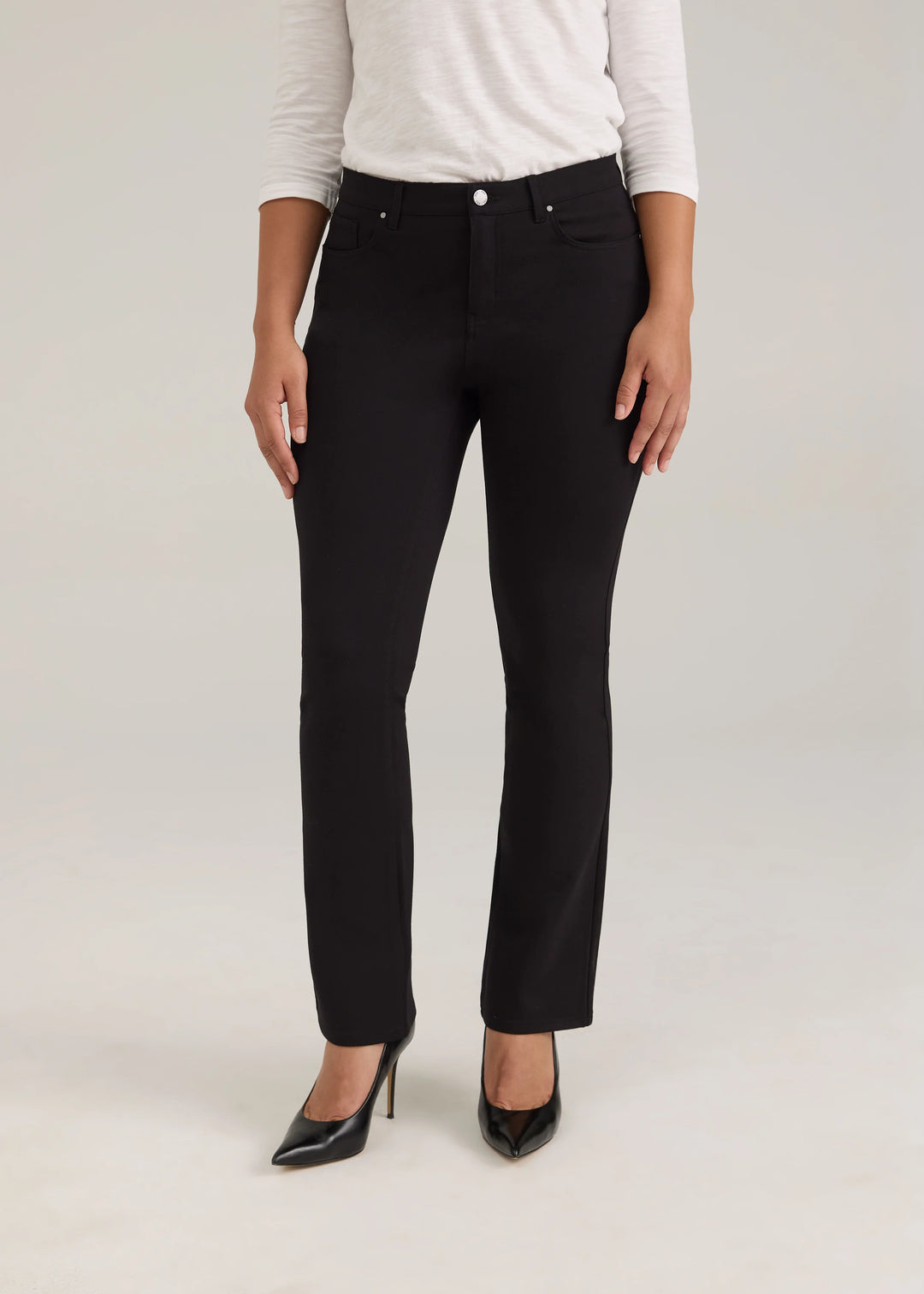 A model wearing a pair of black slim-fit jeans with a white long-sleeve top and black pointed-toe heels, standing against a light gray studio background.