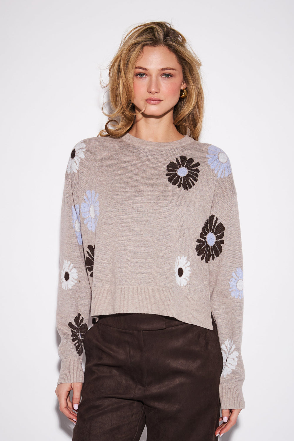 A woman modeling a cropped knit sweater in taupe with large black, white, and pale blue flower motifs, paired with dark brown pants, against a plain light gray studio background.