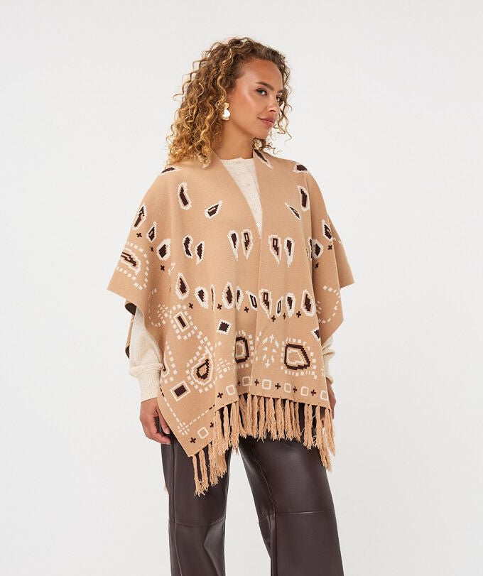 A woman modeling a beige open-front poncho with black and white geometric patterns and fringe, styled with dark brown wide-leg pants and white shoes against a plain white studio background.